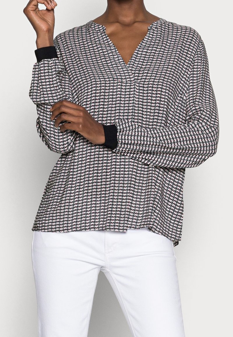 Blouse à motifs noir et blanc avec un col en V, manches longues et poignets noirs, fabriquée dans un tissu léger, associée à un pantalon blanc.