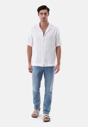 Camicia bianca di lino a maniche corte con chiusura a bottoni, abbinata a jeans azzurri e sandali beige. La texture sembra leggera e traspirante.