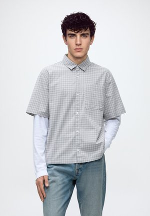 Junger Mann mit lockigem Haar, der ein graues kariertes Kurzarmhemd über einem weißen Langarmhemd und blauen Jeans trägt, steht mit einer Hand in der Tasche.
