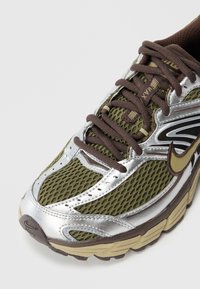 Sapatilha em malha verde oliva com detalhes em sintético prateado, atacadores castanhos e sola texturizada. Apresenta um detalhe do logótipo na lateral.