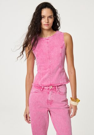 Frau mit langen lockigen Haaren, die ein ärmelloses rosa Denim-Oberteil und passende rosa Denim-Jeans trägt, dazu zwei auffällige Armbänder als Accessoires.