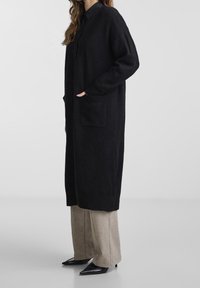 Długi czarny kardigan z miękkiego materiału, z guzikiem z przodu, dwoma przednimi kieszeniami i luźnym krojem. Noszony na beżowe spodnie i czarne buty.