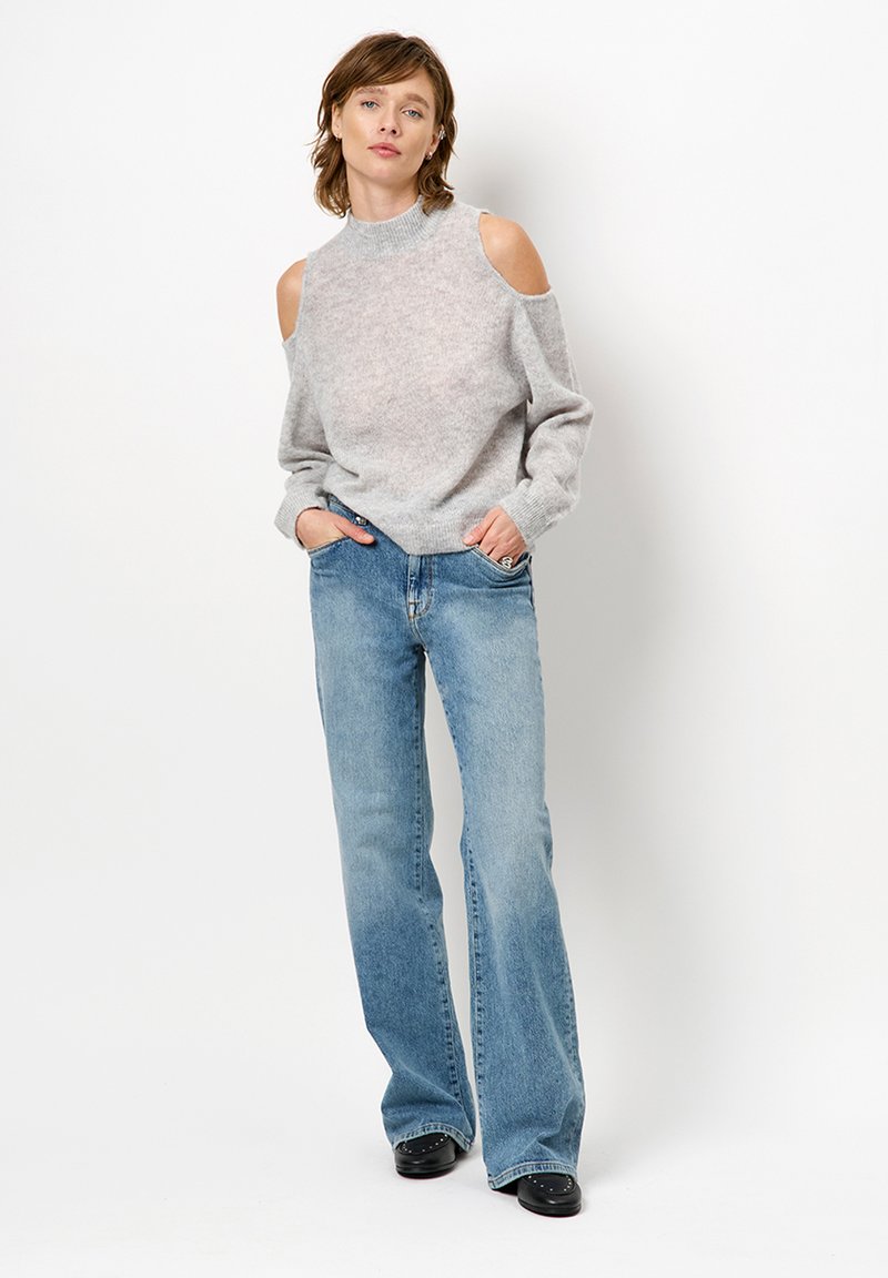 Grau gestrickter Pullover mit offenen Schultern und hohem Kragen, gerippten Bündchen, kombiniert mit hellblauen Schlagjeans, ergänzt durch schwarze Absatzstiefel.