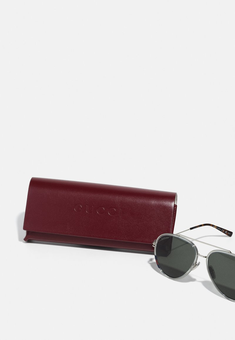 Des lunettes de soleil Gucci dans une monture en métal argenté avec des verres verts, à côté d'un étui en cuir bordeaux embossé du nom de la marque.