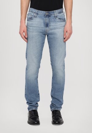 Mann trägt hellblaue Jeans mit geradem Schnitt und schwarze Lederschuhe, steht vor einem schlichten weißen Hintergrund.