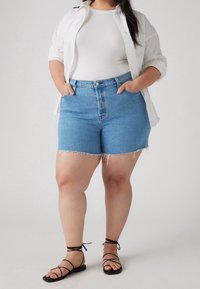 Shorts en denim bleu taille haute avec ourlet effiloché, associés à un débardeur côtelé blanc et une chemise blanche ample à boutons, sandales noires à lanières.