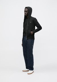 Sweat à capuche noir zippé avec une finition texturée, porté par-dessus un haut noir, associé à un jean bleu foncé et des chaussures blanches. Pose debout, fond neutre.