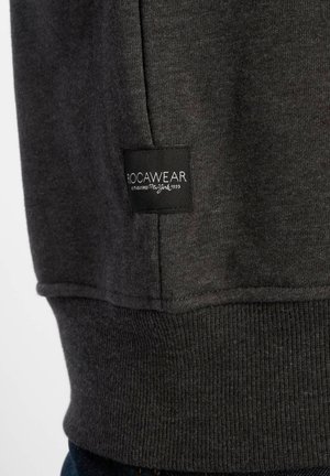 Sötétszürke pulóver bordázott aljjal és mandzsettával. Fekete címke található rajta, rajta a "ROCAWEAR" felirattal és a megalapítás éve: 1999. Puha tapintású.