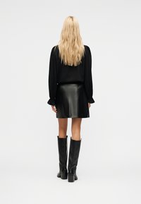 Γυναίκα με μακριά ξανθά μαλλιά που φορά μαύρο πουκάμισο, κοντή μαύρη δερμάτινη φούστα και μαύρες μπότες μέχρι το γόνατο, γυρισμένη με την πλάτη προς την κάμερα, σε λευκό φόντο.