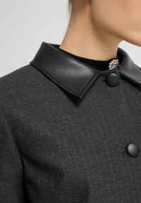 Czarny płaszcz z mieszanki wełny z błyszczącym kołnierzem, dużymi czarnymi guzikami oraz subtelnym wzorem w prążki. Charakteryzuje się teksturowanym materiałem.