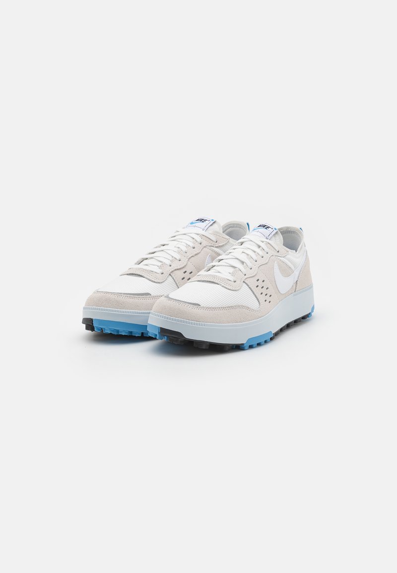 Zapatillas beige y blancas con material de ante y malla, que cuentan con una suela de goma azul, acentos perforados y el logo de Nike en blanco.