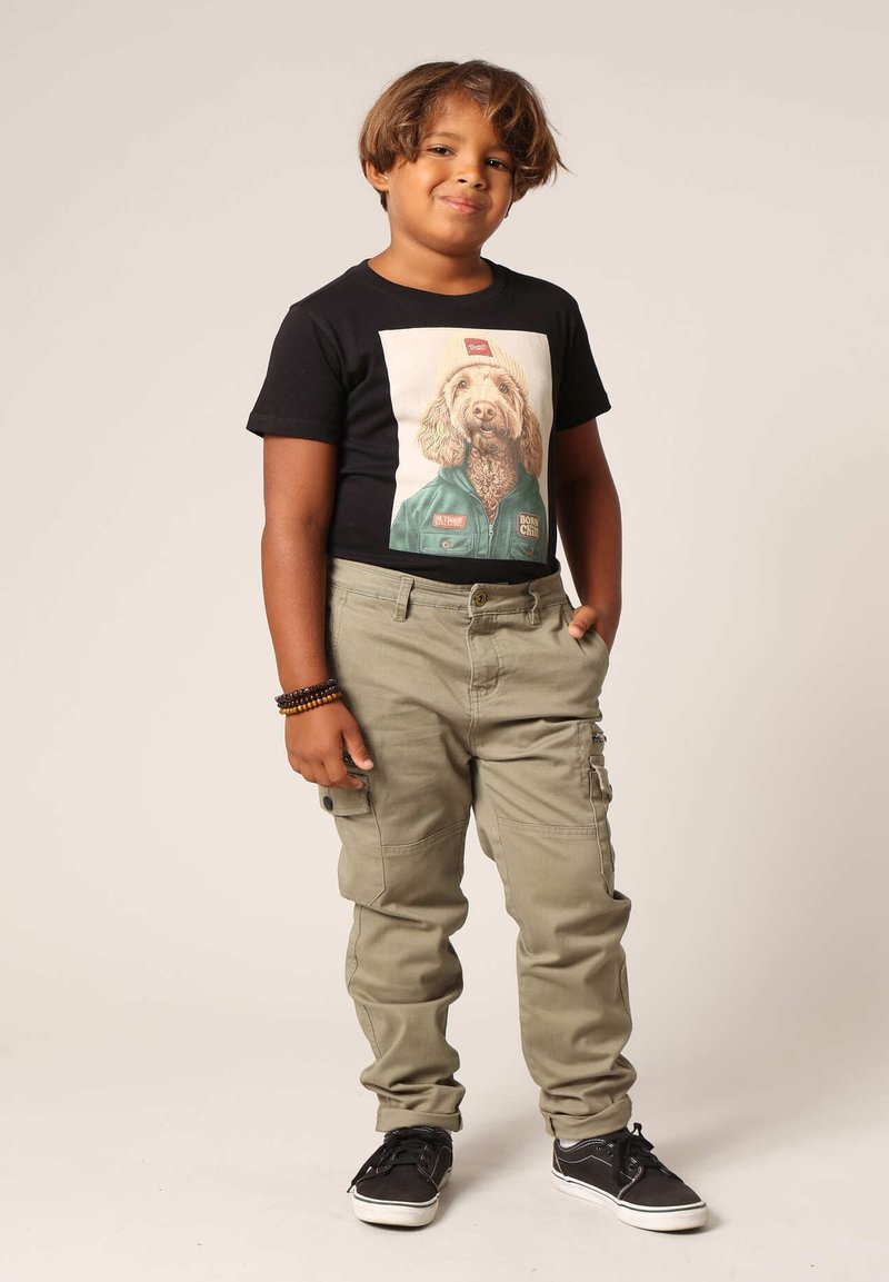 Zwarte T-shirt met een hondenafbeelding; khaki cargo broek met zakken; zwarte schoenen; polsband; lichtbruin haar. Neutrale achtergrond.