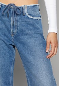 Ljusblå denimjeans med fransad midja och dragsko. Funktioner inkluderar standardfickor och en avslappnad passform. Strukturerad yta.