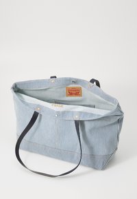 Striped tote bag with blue and white fabric, black handles, and gold hardware. Interior features a red label with brand logo and stitching accents.