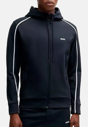 Uomo che indossa una felpa nera con zip con piping bianco sulle maniche e logo BOSS sul petto e sui pantaloni, su sfondo semplice.