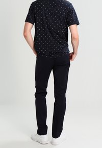 Námořnická košile s krátkým rukávem s bílými puntíky, zkombinovaná s tmavými kalhotami a bílými teniskami. Model zobrazen zezadu, obrácený vpravo.