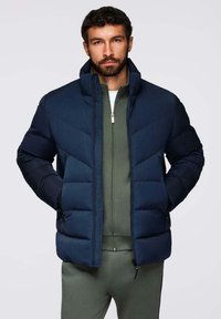 Giacca a vento blu navy con design a zigzag, dotata di alto colletto, chiusura con zip frontale e due tasche laterali. Indossata sopra una felpa verde con zip.