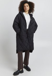 Manteau long noir matelassé avec capuche, doté de fermetures à boutons et d'une coupe ample, porté sur un pantalon marron et des chaussures à semelle épaisse noires.