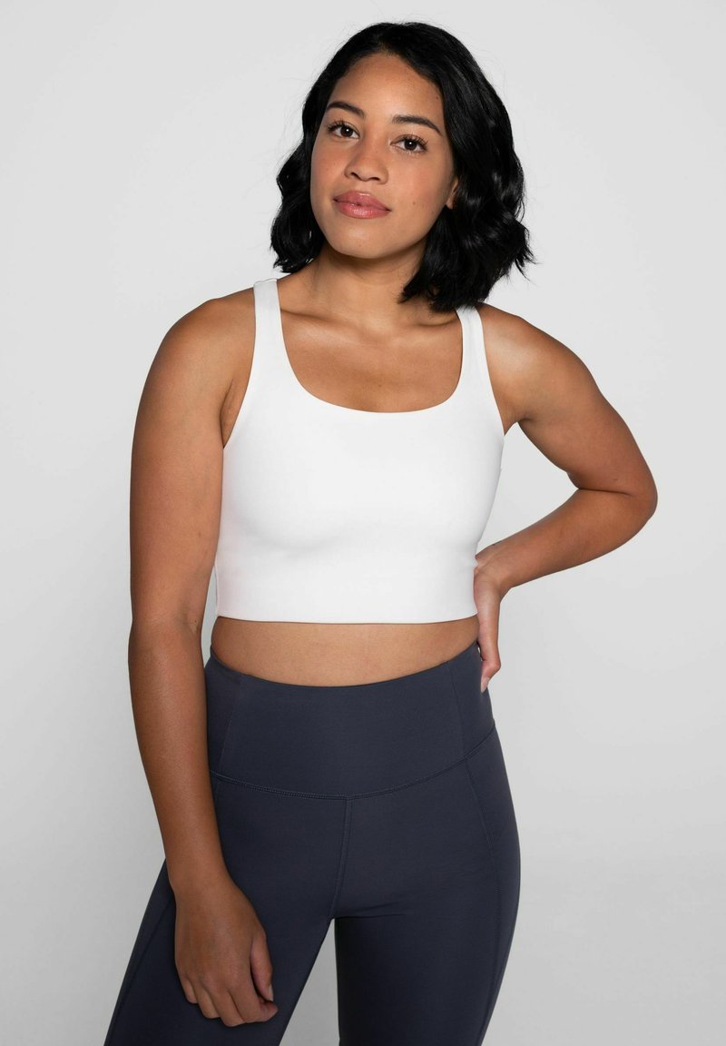 Top corto blanco con escote cuadrado y tirantes anchos, combinado con leggings oscuros. Textura suave, diseño ajustado y sin patrones visibles.
