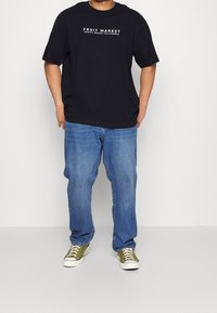 T-shirt nera in cotone con la scritta "FRUIT MARKET VENICE BEACH CALIFORNIA", abbinata a jeans blu e sneakers verde oliva.