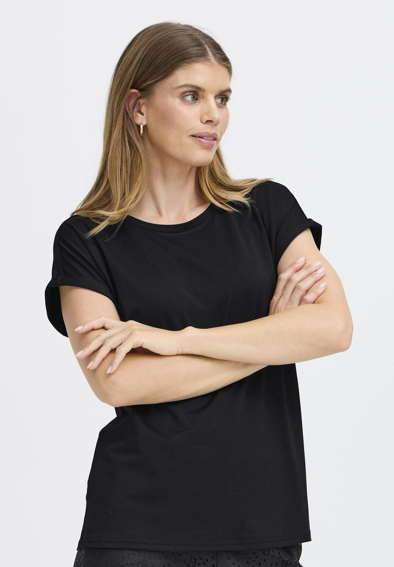 Femme aux longs cheveux blonds portant des boucles d'oreilles en créoles dorées et un t-shirt noir à manches courtes, bras croisés, regardant sur le côté sur un fond clair.