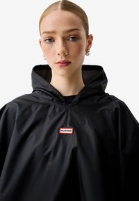 Νεαρή γυναίκα με τα μαλλιά κομμένα πίσω, φορά σκουλαρίκια με πέρλες και μαύρο αδιάβροχο πόντσο με κουκούλα και λογότυπο Hunter, μπροστά σε λευκό φόντο.