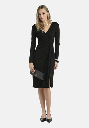 Black wrap dress with long sleeves, featuring a front slit, textured fabric, and a streamlined silhouette. Paired with a black clutch.