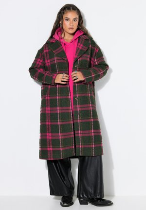 Mujer con un abrigo largo de cuadros en verde oscuro y rosa sobre una sudadera con capucha rosa brillante y pantalones negros de pierna ancha con zapatos negros.