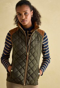 Olivovo zelená prešívaná vesta s hnedými semišovými ramenami, dvoma bočnými vreckami a predným zipsom. Nosená cez námořnícku a béžovú pruhovanú košeľu.