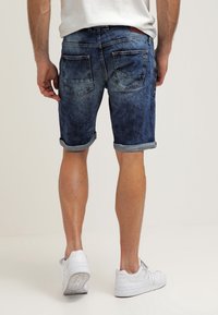 Shorts in denim di un blu scuro, leggermente sbiaditi, con risvolti e due tasche posteriori. Indossati con sneakers bianche su uno sfondo neutro.