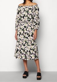 Schulterfreies Kleid mit schwarzem Hintergrund und floralem Druck in weißen Lilien. Elastischer Taillenbund und rüschenbesäumter Saum, kombiniert mit schwarzen Plateausandalen.