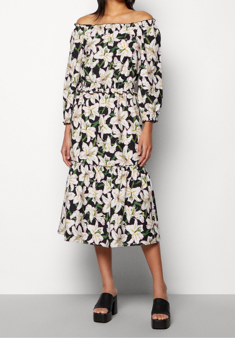 Schulterfreies Kleid mit schwarzem Hintergrund und floralem Druck in weißen Lilien. Elastischer Taillenbund und rüschenbesäumter Saum, kombiniert mit schwarzen Plateausandalen.