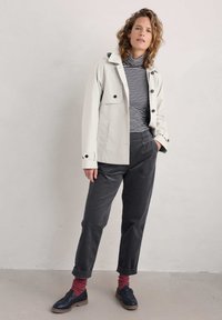 Šviesiai smėlinė vandeniui atspari striukė su juodomis mygtukais, derinama su tamsiai mėlynu ir baltu dryžuotu polo megztiniu, pilkais kostiumo kelnais ir tamsiais batais.