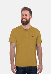 Senfgelbes, geripptes Kurzarmshirt mit drei Knöpfen und schwarzem Bärenlogo auf der linken Brust. Getragen mit dunklen Jeans.