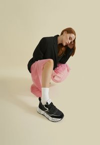 Sneaker bianco e nero con un logo audace sulla suola, materiali in suede e mesh, abbinato a calzini bianchi e una gonna plissettata rosa.