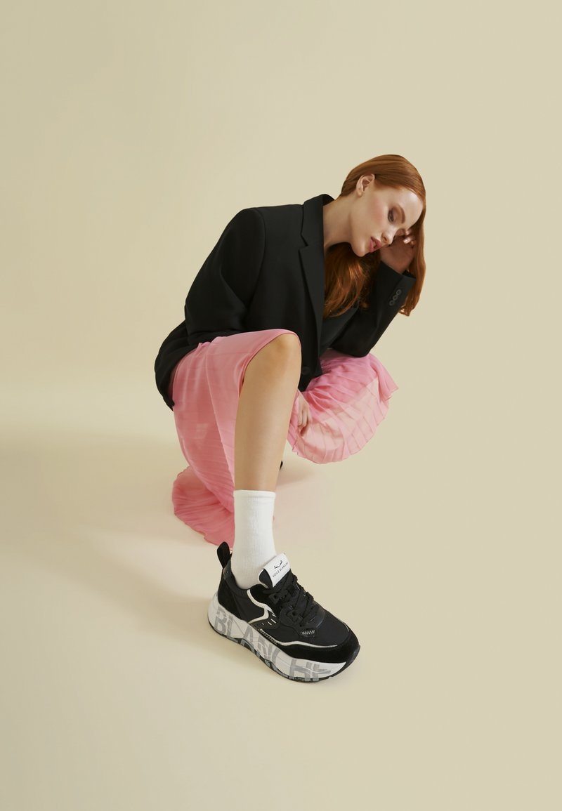 Sneaker bianco e nero con un logo audace sulla suola, materiali in suede e mesh, abbinato a calzini bianchi e una gonna plissettata rosa.
