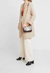 Cardigan en tricot beige, chemisier rouge rouille, pantalon en velours côtelé crème à jambes larges, bottes à talons noires et un sac à main à motif serpent avec quincaillerie dorée.