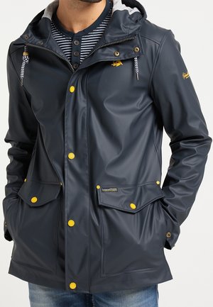 Homme portant une veste imperméable bleu marine avec des boutons jaunes et une capuche, les mains dans les poches, par-dessus une chemise henley à rayures bleues et un jean.