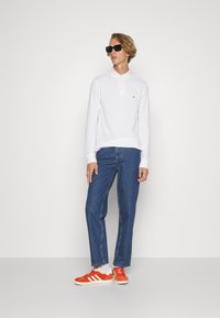 Hvit langermet polo skjorte med krage, blå jeans, og oransje joggesko med hvite striper. Modellen har på seg svarte solbriller.