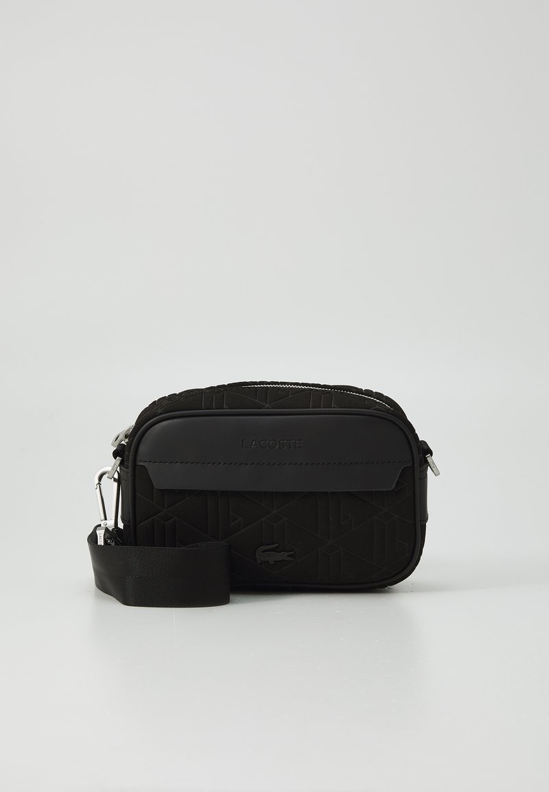 Sac à bandoulière Lacoste noir avec un motif géométrique texturé, un patch logo lisse, une fermeture zippée et une bandoulière réglable. Forme rectangulaire compacte.