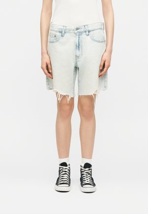 Isik, kes kannab helesiniseid kulunud teksapükse, valgeid sokke ja musti kõrge säärega tossusid, seisab valge tausta ees.