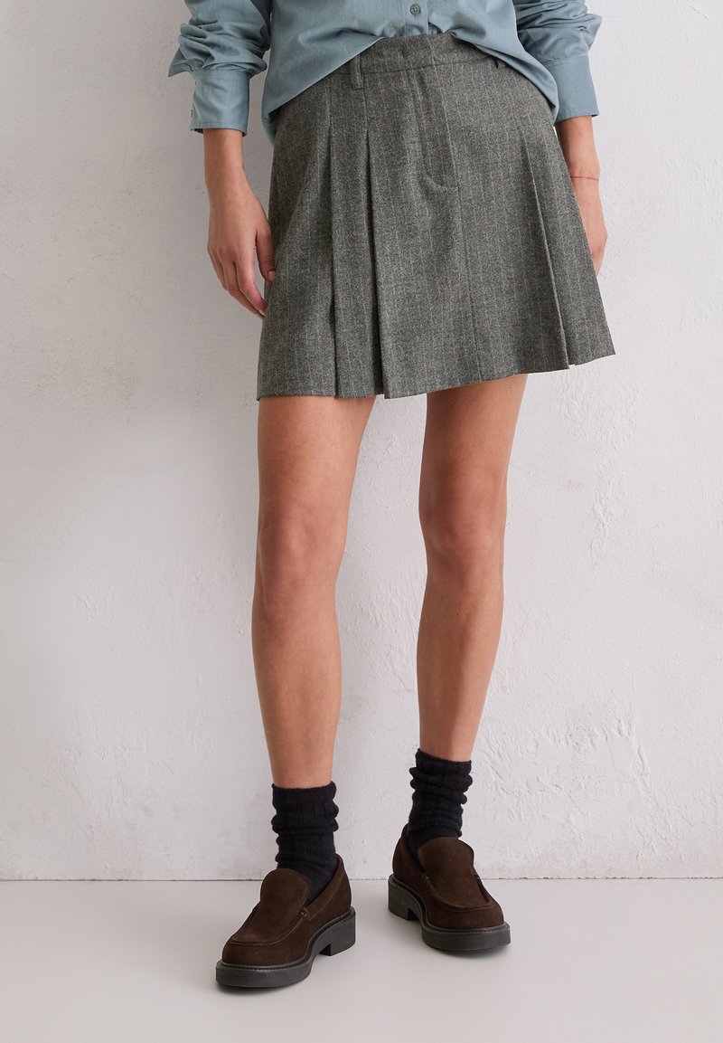 Grijze geplooide rok van gestructureerde stof, gecombineerd met donkerbruine suède schoenen en zwarte geribbelde kousen. De outfit heeft een blauwe shirt.
