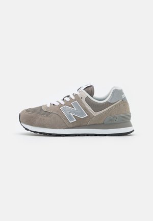 Widok z boku szarego buta sportowego New Balance z panelami z zamszu i siateczki, białymi sznurówkami oraz białą i czarną podeszwą na jasnym tle.