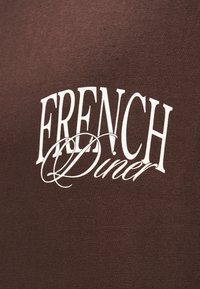 Καφέ ύφασμα με λευκό κείμενο που γράφει "FRENCH Diner." Το κείμενο συνδυάζει γράμματα με πατούρες και καλλιγραφικά γράμματα, δημιουργώντας μια τολμηρή αντίθεση.