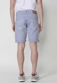 Vīrietis valkā gaiši zilus sarullētus denim šortus, baltu T-kreklu un baltas tenisa kurpes ar sarkaniem, ziliem un pelēkiem akcentiem, stāvot pret vienkāršu fonu.