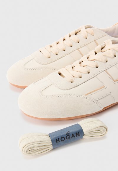 Para beżowych sneakersów z zamszowej i skórzanej powierzchni z dopasowanymi sznurówkami oraz dodatkowym zestawem beżowych sznurówek oznaczonych "HOGAN" na białym tle.