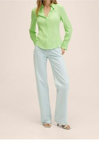 Lichtgroene blouse met lange mouwen en knoopsluiting, gecombineerd met lichtblauwe, wijde jeans. Witte sandalen met enkelbandje.