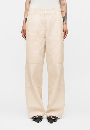 Μπεζ παντελόνια με φαρδιά μπατζάκια, μεγάλες μπροστινές τσέπες με πατ και λεπτομέρειες με κουμπιά, φορεμένα με μαύρα παπούτσια με μυτερή μύτη και ασημένιες αγκράφες.