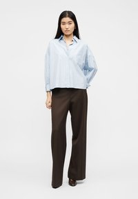 Γυναίκα στέκεται φορώντας μια χαλαρή ανοιχτό μπλε πουκαμίσα με ρίγες και κουμπιά, σκούρο καφέ φαρδιά παντελόνια και καφέ παπούτσια σε απλό λευκό φόντο.