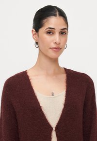 Burgundský pletený cardigan s jemnou texturou, otevřeným předním dílem a jedním knoflíkem na zapínání. Nosič má na sobě lehký, třpytivý krémový top.